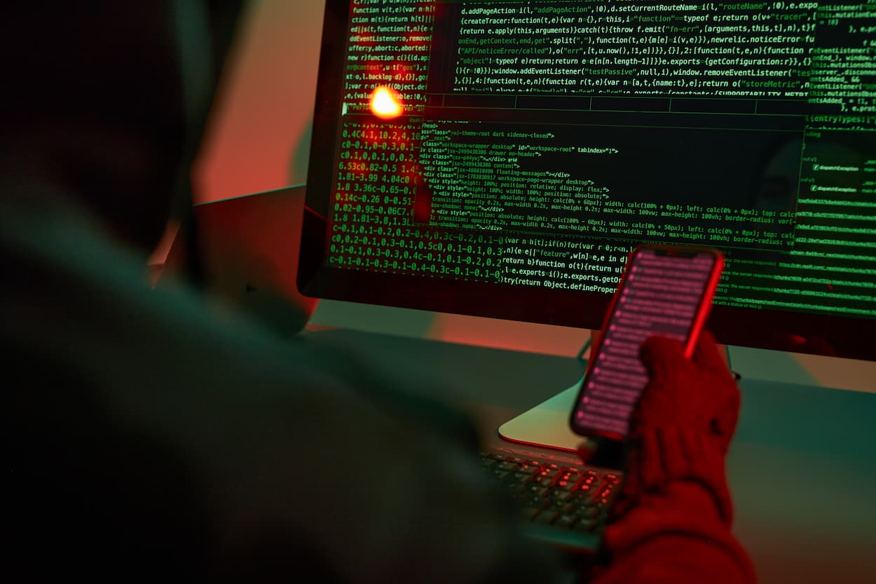 Persona con capucha frente a una pantalla de computadora que muestra código, representando un posible ataque de phishing.