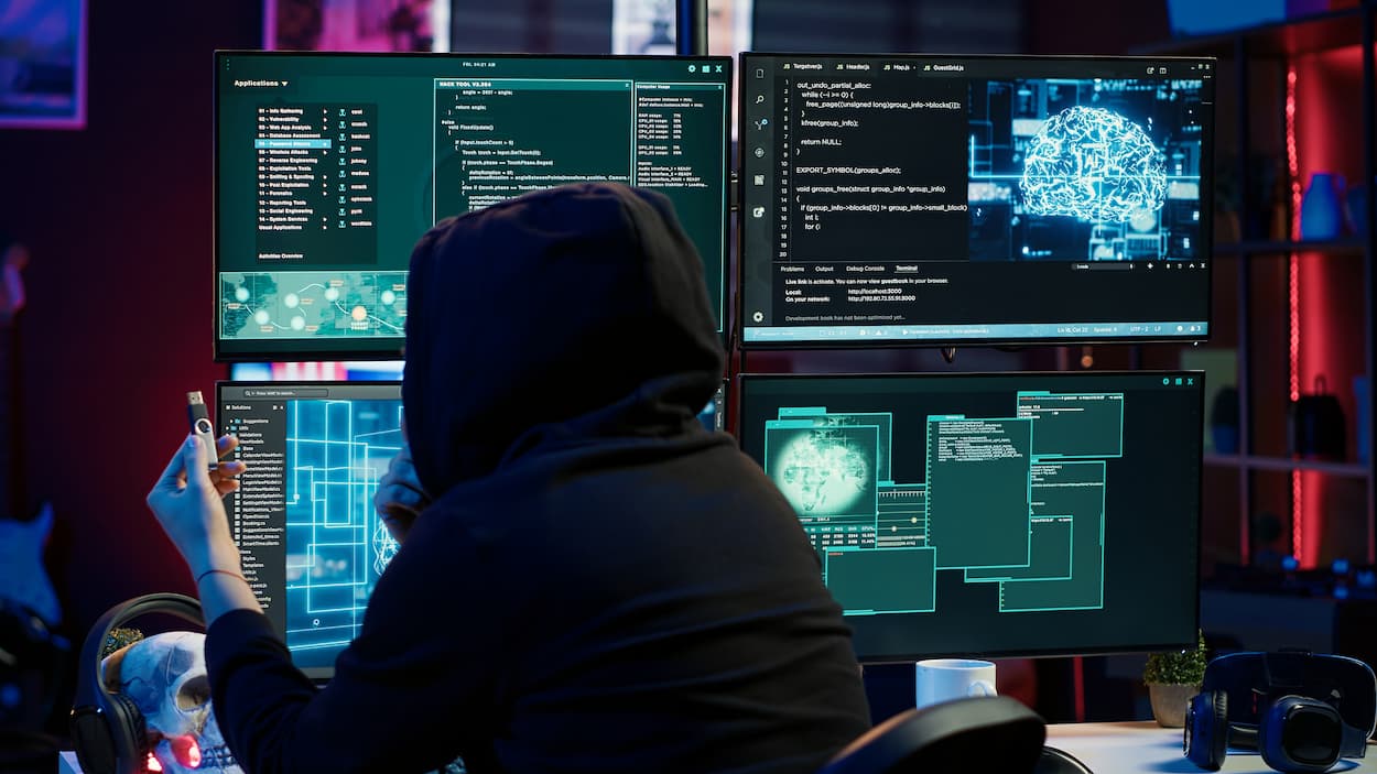 Persona con capucha frente a un ordenador con múltiples pantallas, representando un posible ataque de phishing.